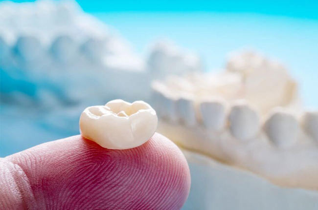 White dental crown held on fingertip with model teeth and blue sky background