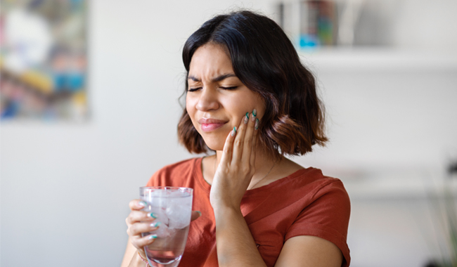Woman holding water glass, touching cheek in pain, experiencing tooth sensitivity or dental discomfort indoors.