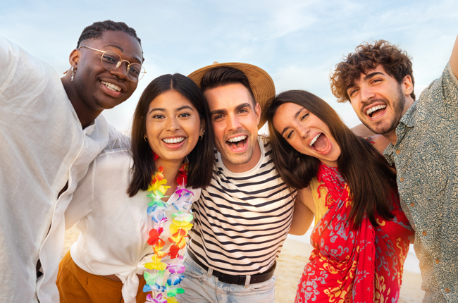 Five diverse young adults smiling together outdoors against a bright sky.