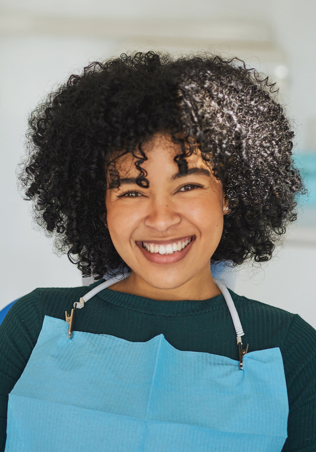 Young woman with curly black hair smiling warmly while wearing a blue apron in a dental office setting.