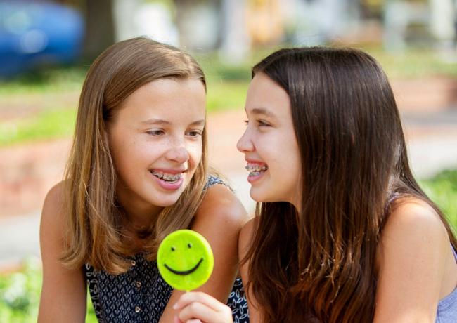 Two smiling girls holding a green smiley face lollipop outdoors in a park setting.